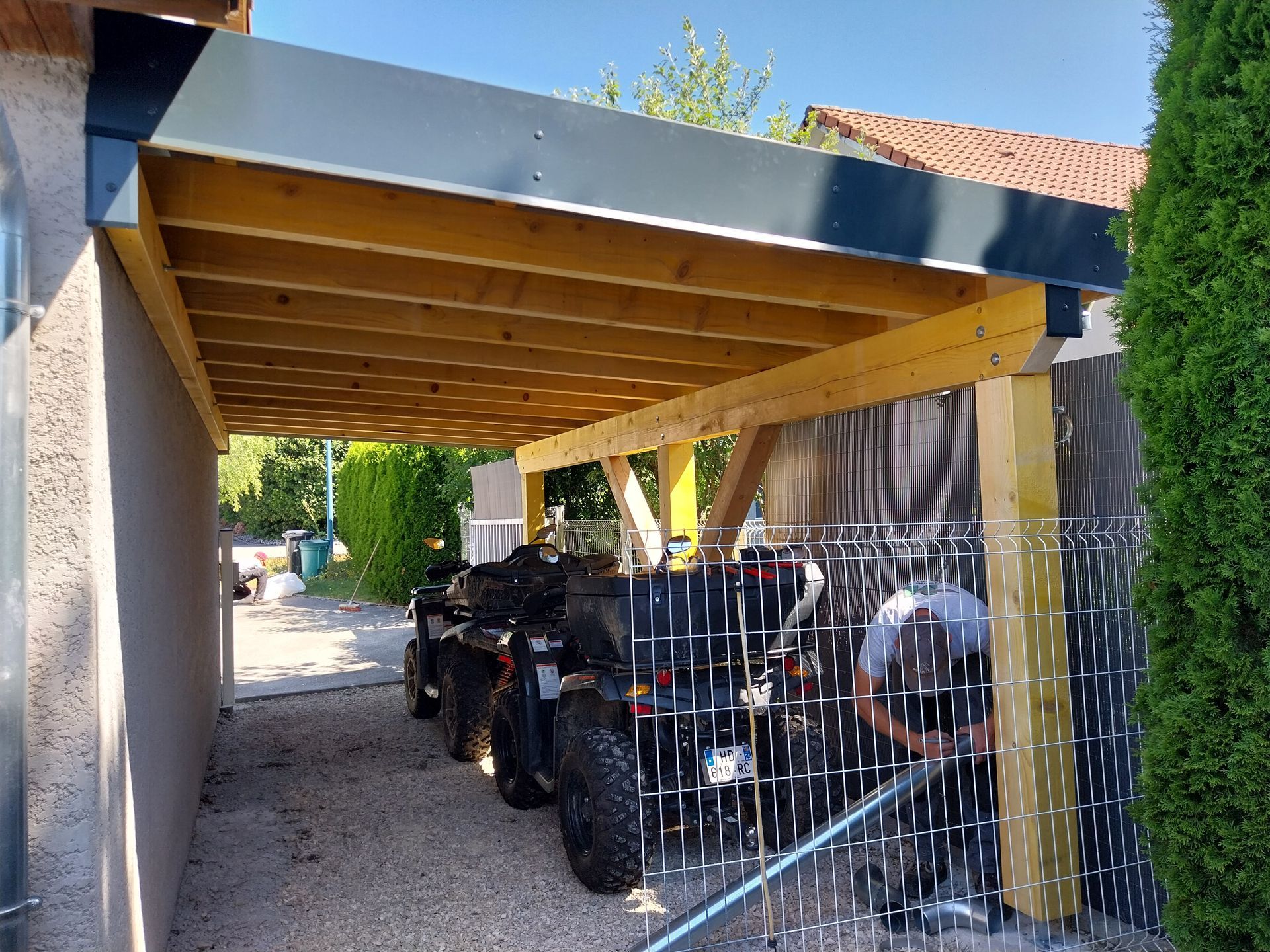 Abri en bois pour quads, attenant à une maison. Toit noir, supports en bois beige, panneau latéral gris.