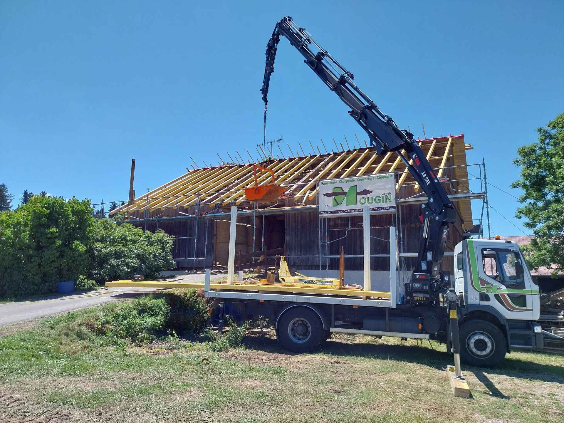 Un camion-grue soulève des matériaux de construction pour les déposer sur le toit d'un immeuble sous un ciel bleu azur.
