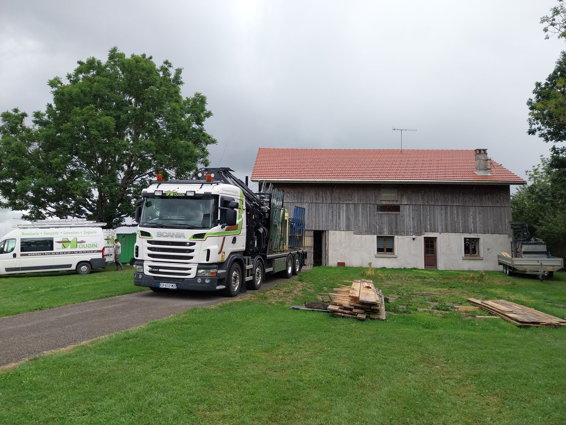 Un camion-grue est stationné devant un bâtiment en bois délavé par les intempéries, au toit de tuiles rouges.