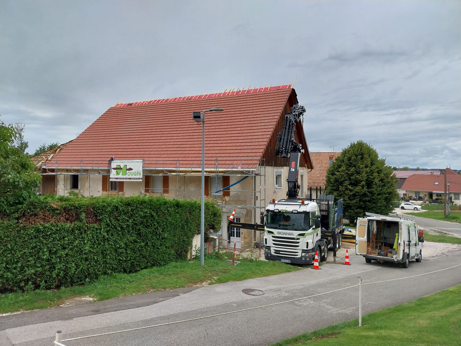 Une grue hisse des matériaux sur le toit de tuiles rouges d'un bâtiment, tandis qu'un camion et une camionnette blancs sont garés à proximité.