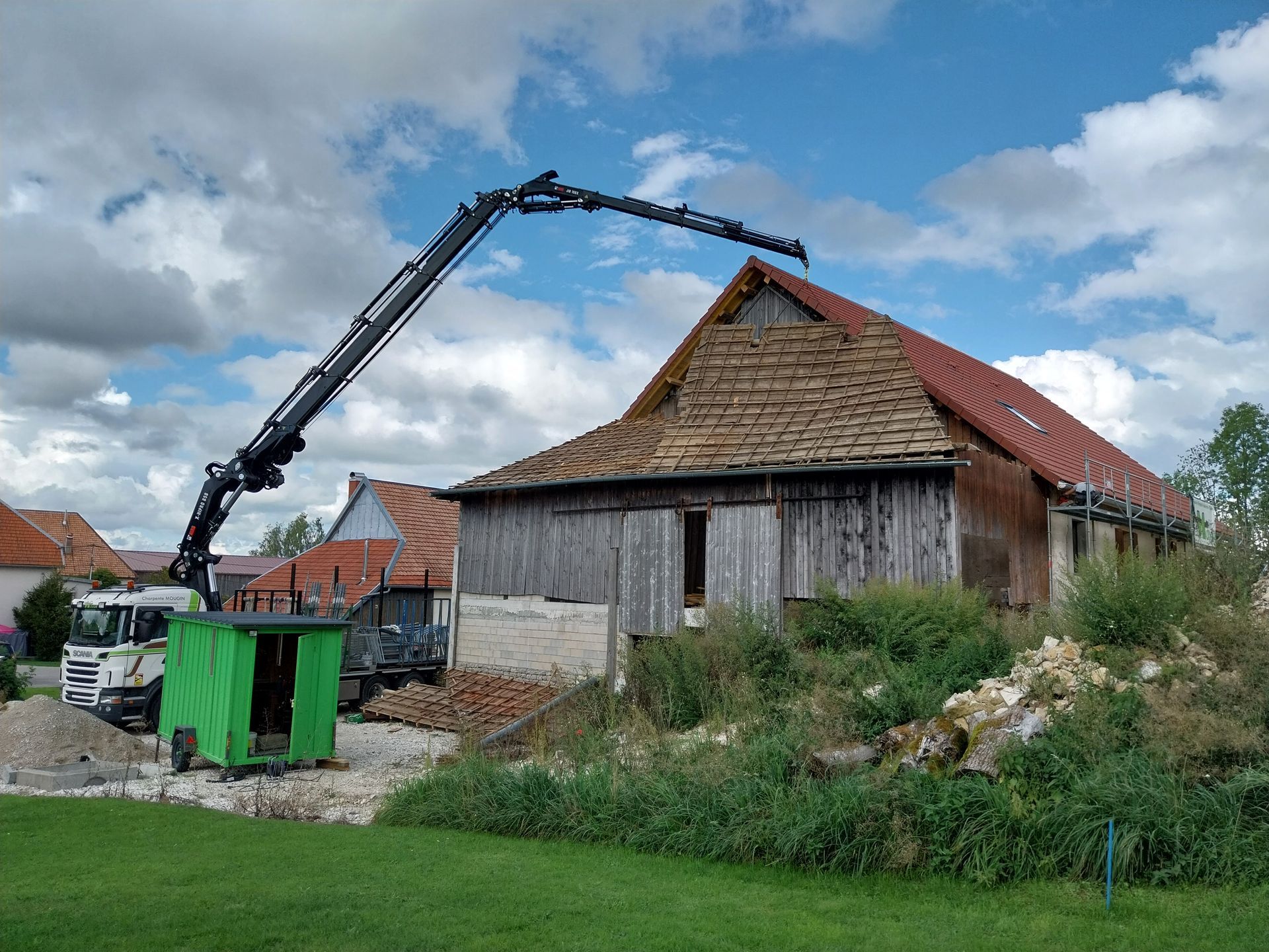Une grue démolit une grange délabrée au toit rouge, un hangar vert à proximité et un ciel bleu en arrière-plan.