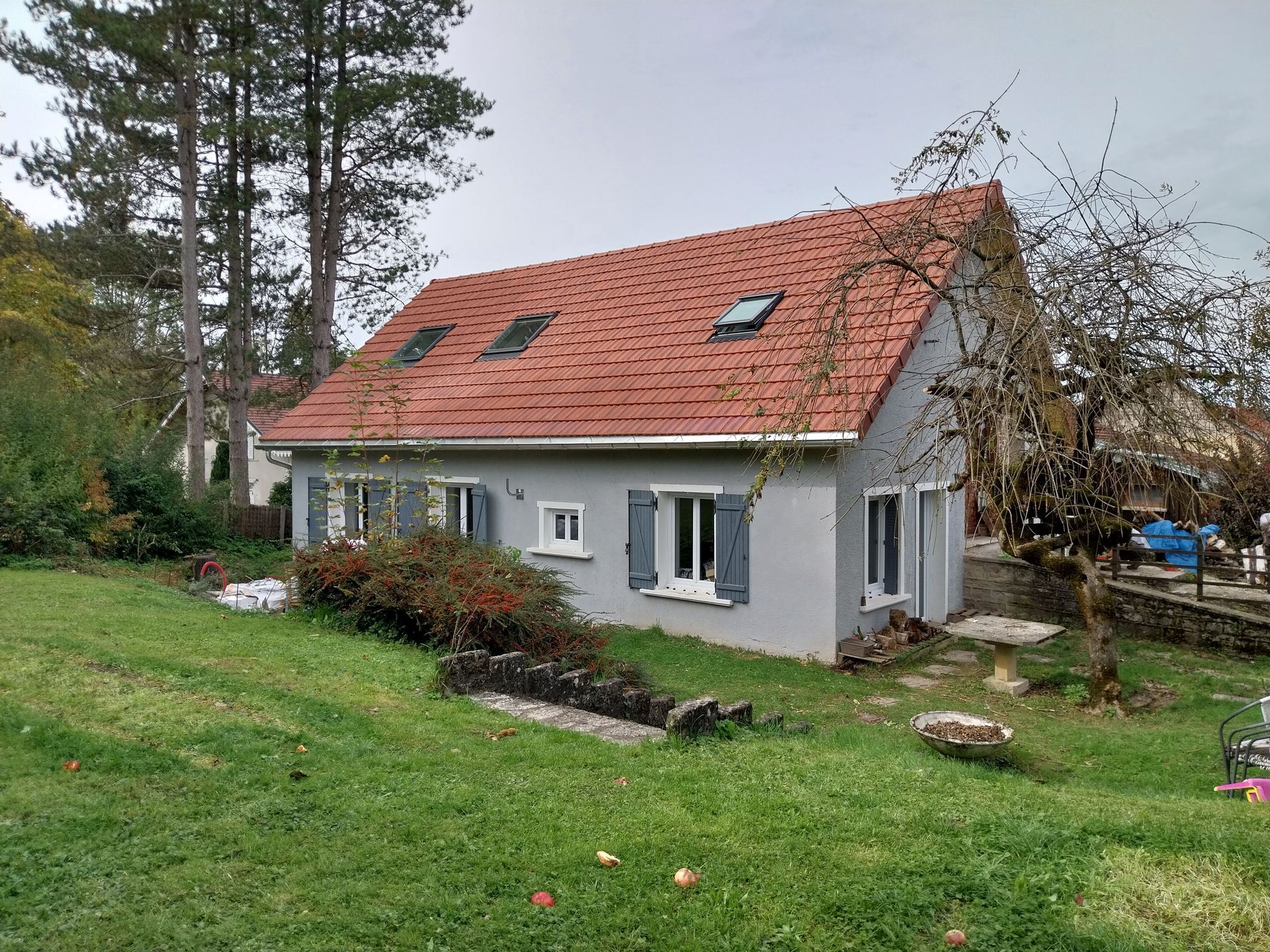Maison grise au toit rouge sur une colline herbeuse, avec des buissons et des arbres en arrière-plan.