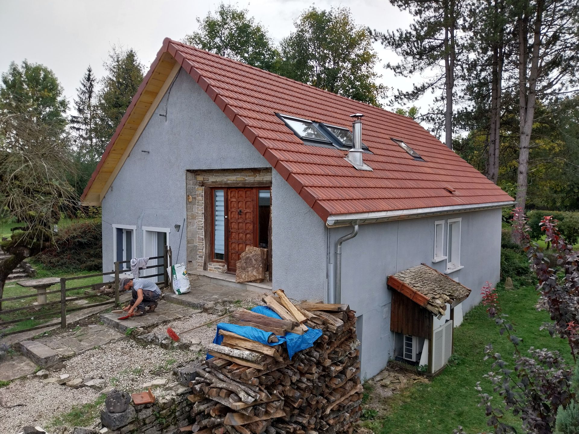 Petite maison au toit de tuiles rouges, murs gris, porte ouverte, pile de bois devant.