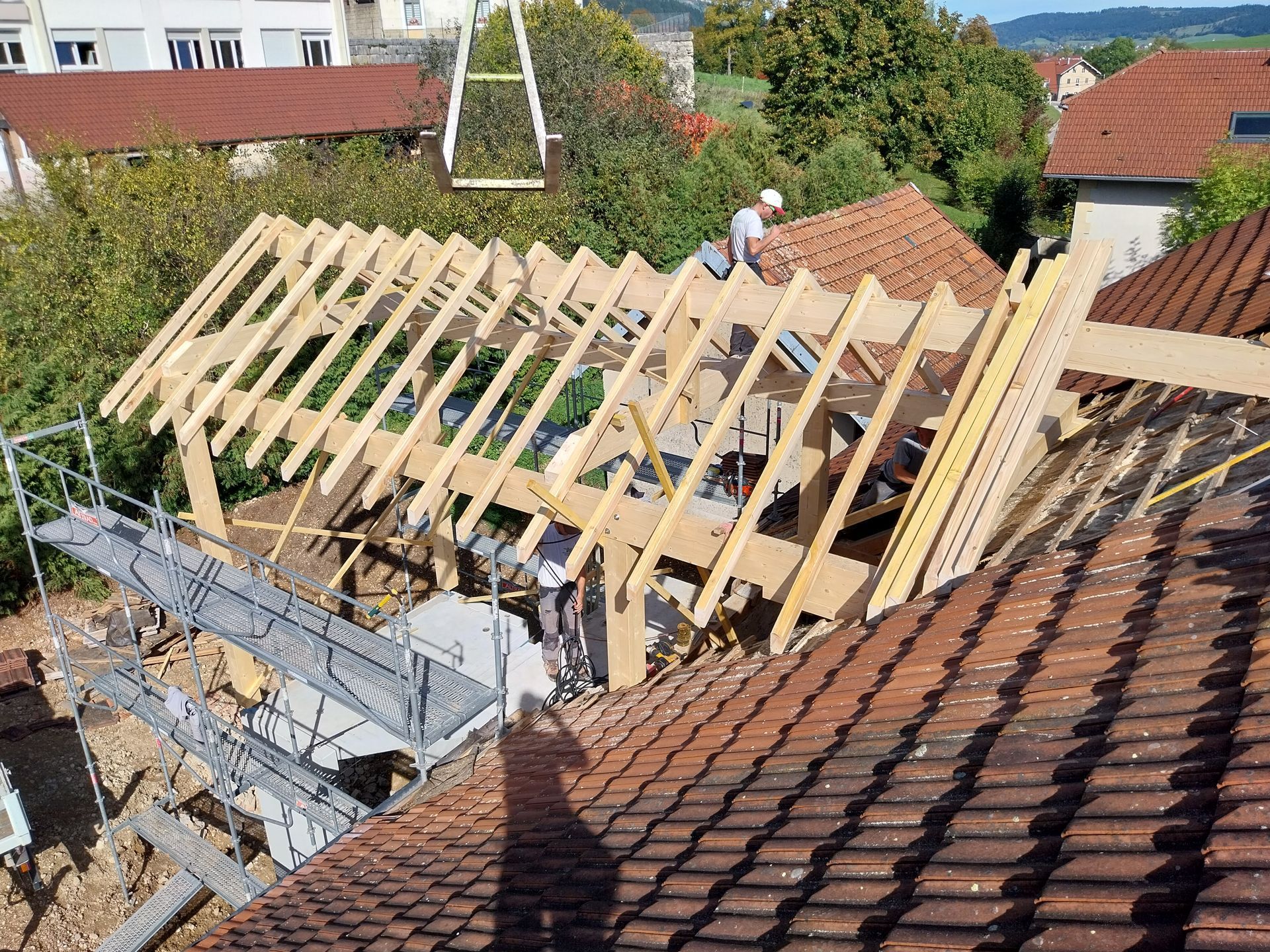 Des ouvriers du bâtiment construisent une charpente de toit en bois sur un toit de tuiles ; échafaudages et grue visibles.