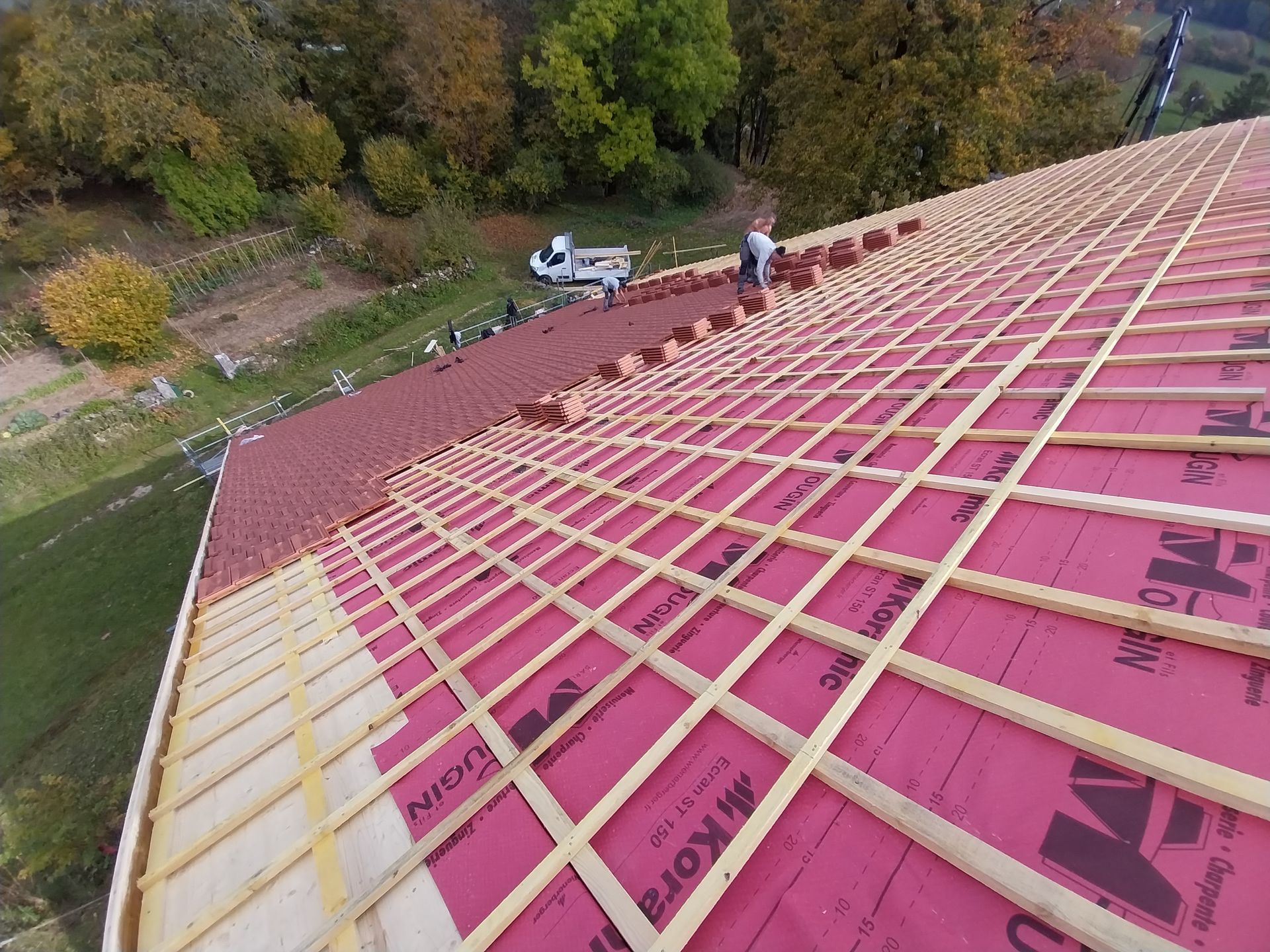 Des ouvriers du bâtiment posent des tuiles rouges sur un toit. Charpente et sous-toiture en bois visibles. En extérieur, en journée.