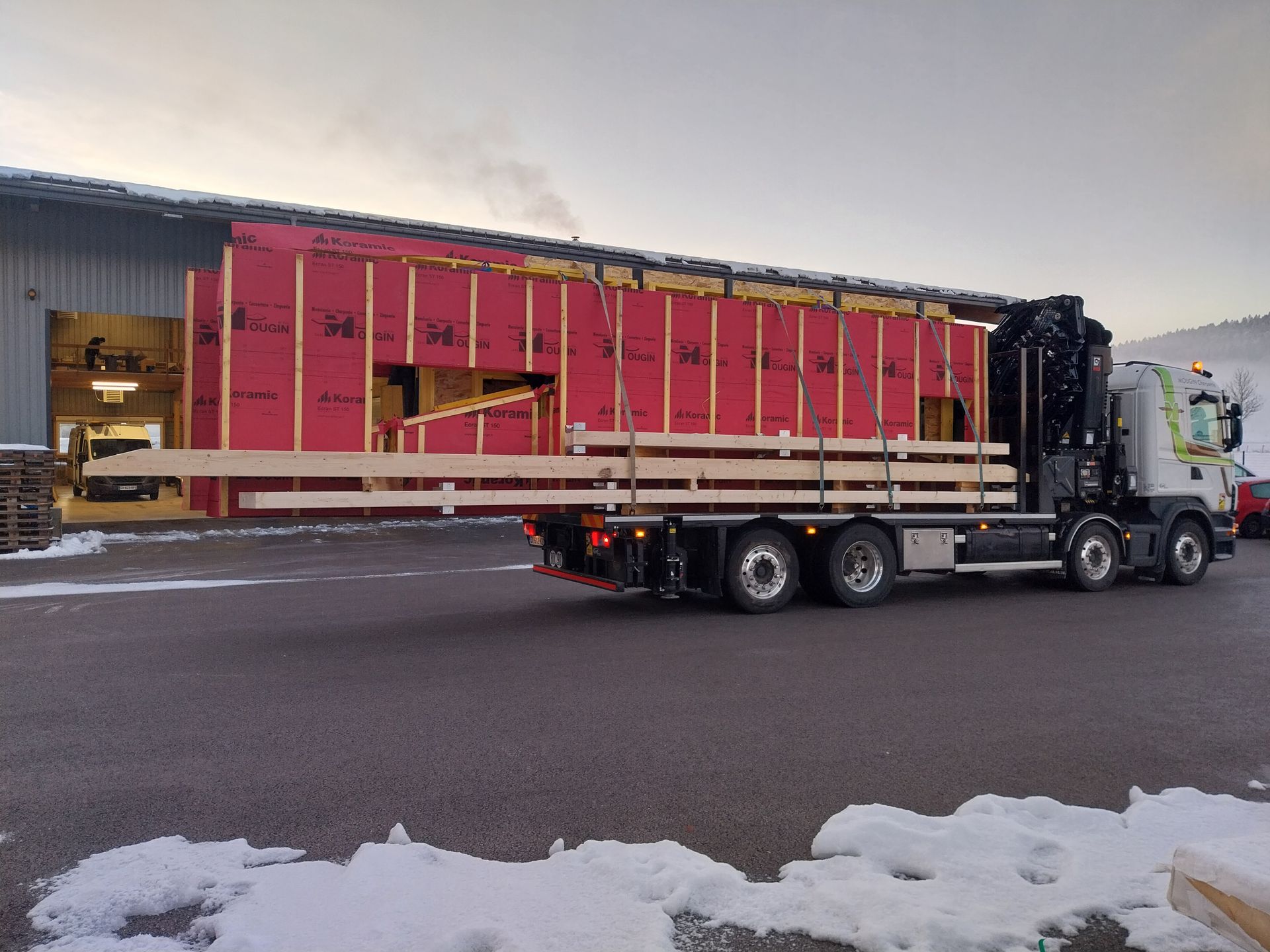 Un camion chargé d'éléments de construction en bois est en cours de chargement près d'un bâtiment au revêtement rouge.