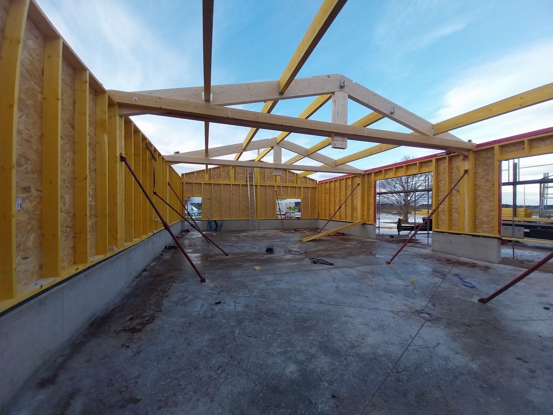 Vue intérieure d'un bâtiment en construction : charpente en bois, poutres et poteaux de soutien. Murs jaune vif, ciel bleu.