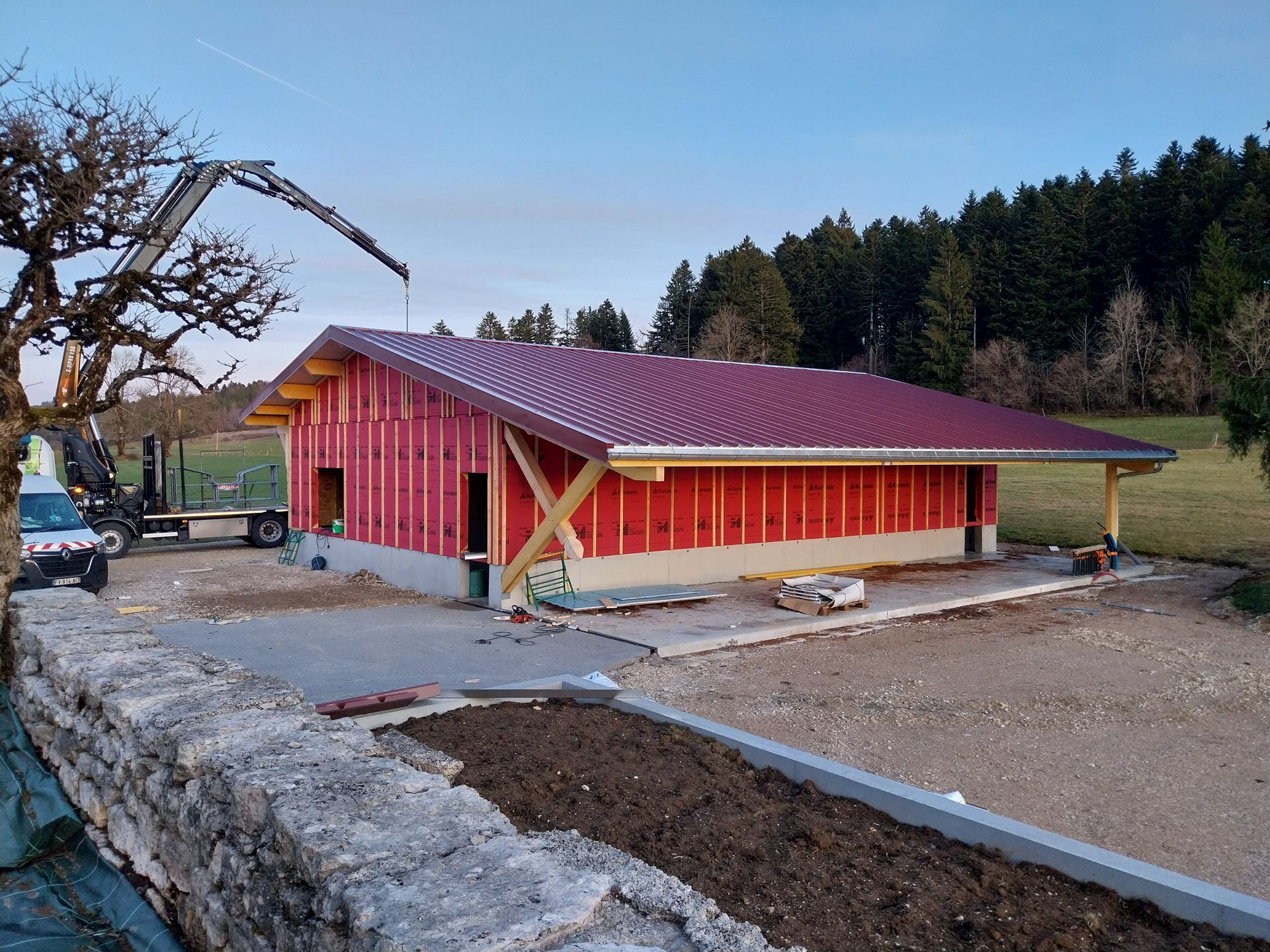 Bâtiment en construction, toit rouge et ossature bois. Fondations en béton, entouré de verdure.