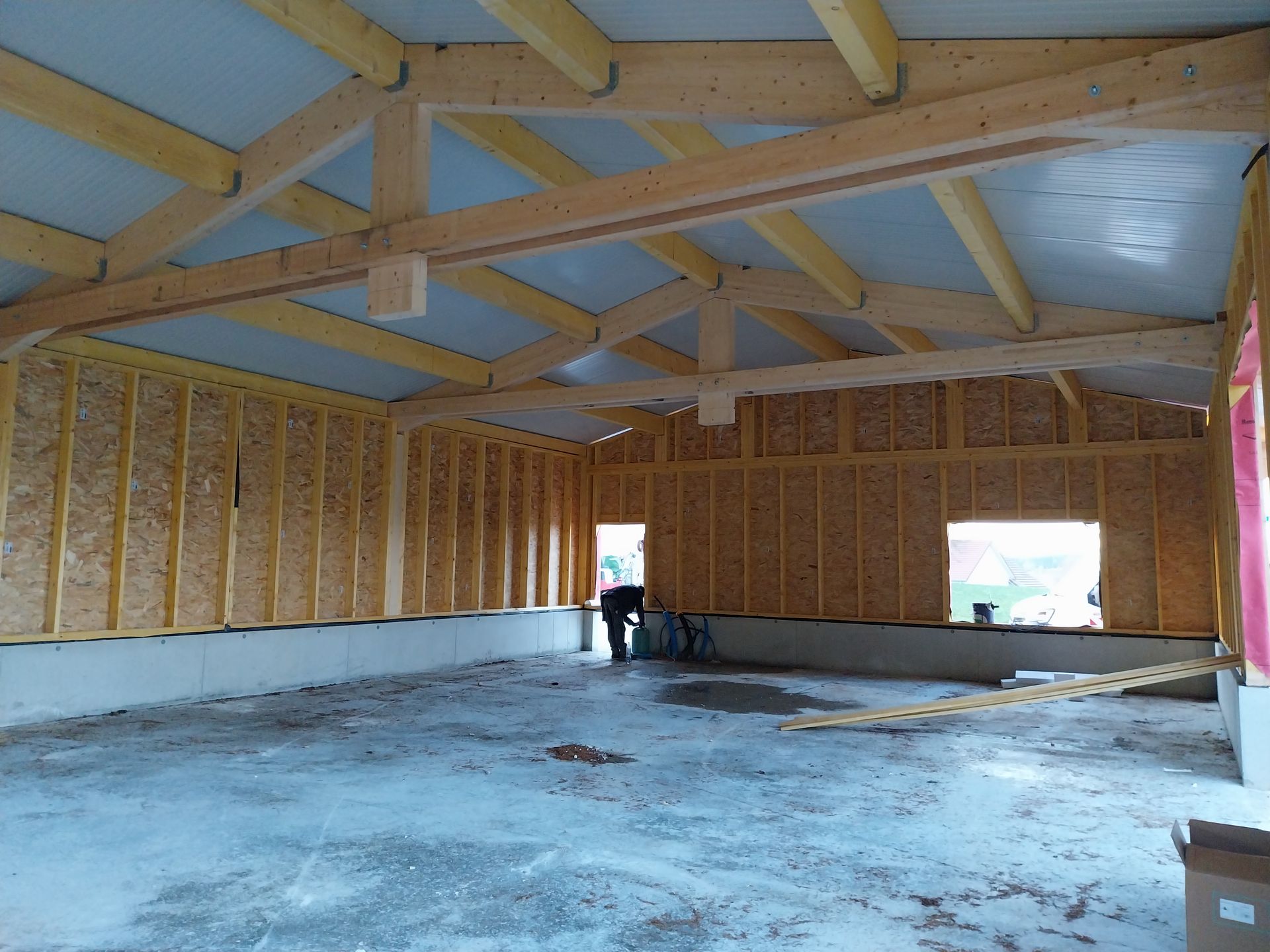 Intérieur d'un bâtiment en construction, montrant des poutres en bois, des murs, un sol en béton et une personne au travail.