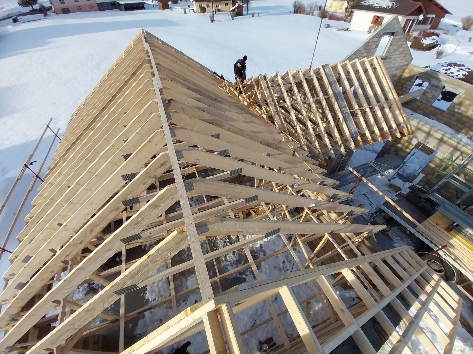 Construction d'une charpente de toit en bois dans un décor enneigé, un ouvrier sur le faîte.