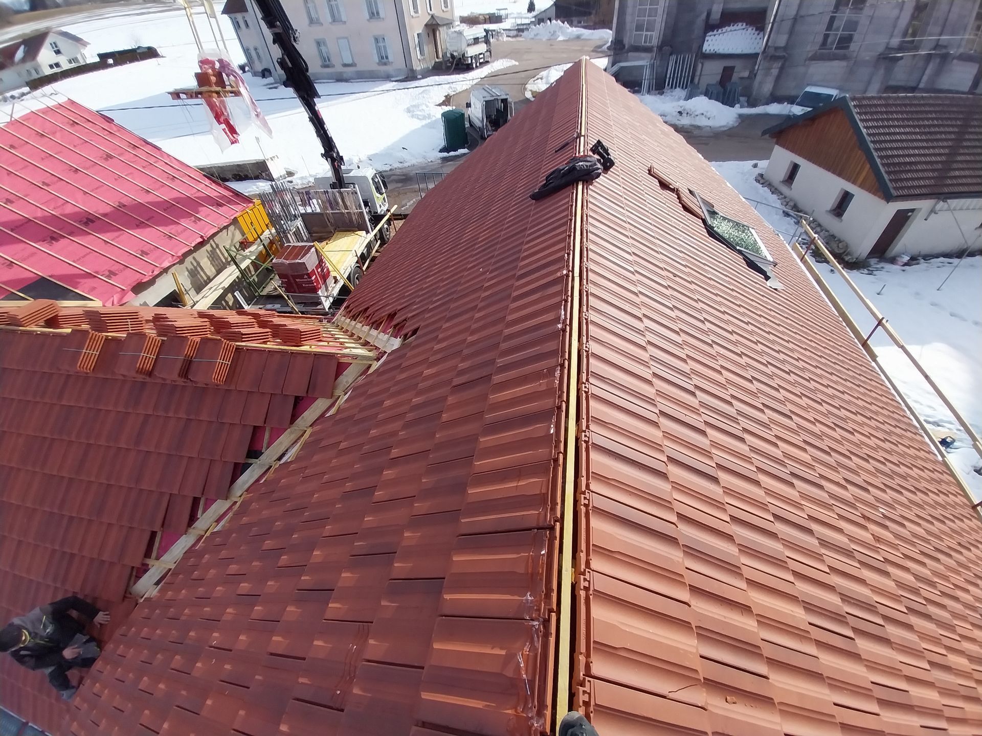 Un couvreur travaille sur un toit de tuiles rouges dans un décor enneigé, avec une grue en arrière-plan.