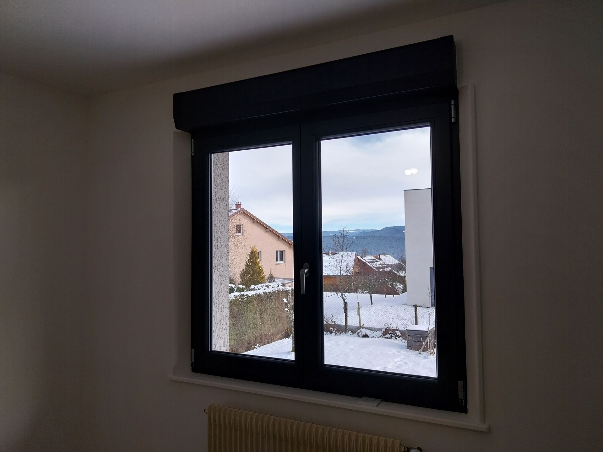 Fenêtre avec store enrouleur noir fermé, donnant sur un village enneigé et des montagnes.