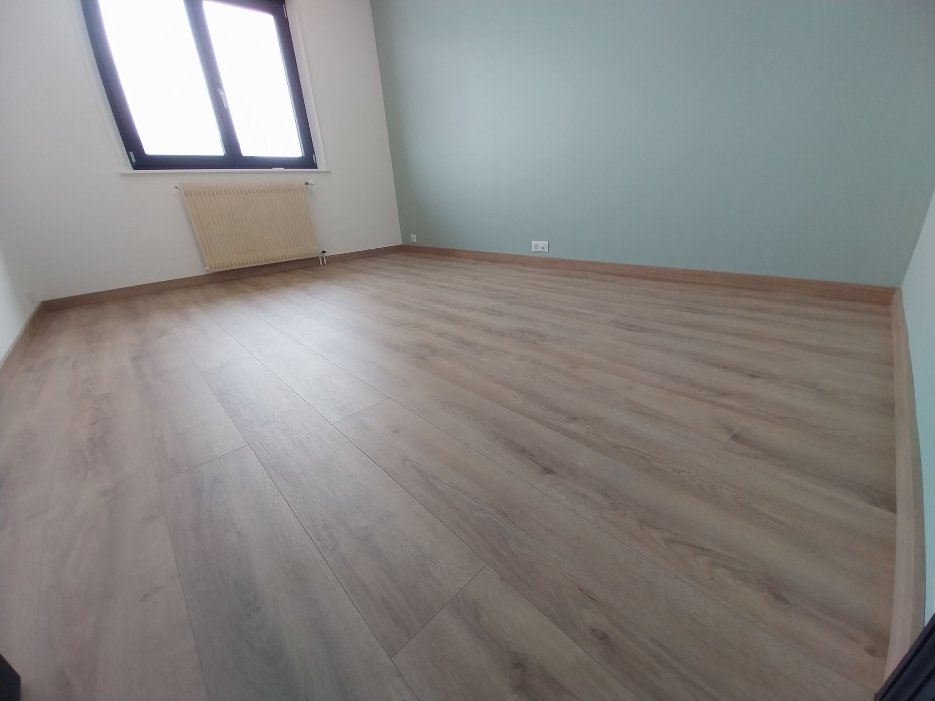 Pièce vide avec un sol clair imitation bois, une fenêtre, un radiateur et des murs bicolores : blanc et turquoise.