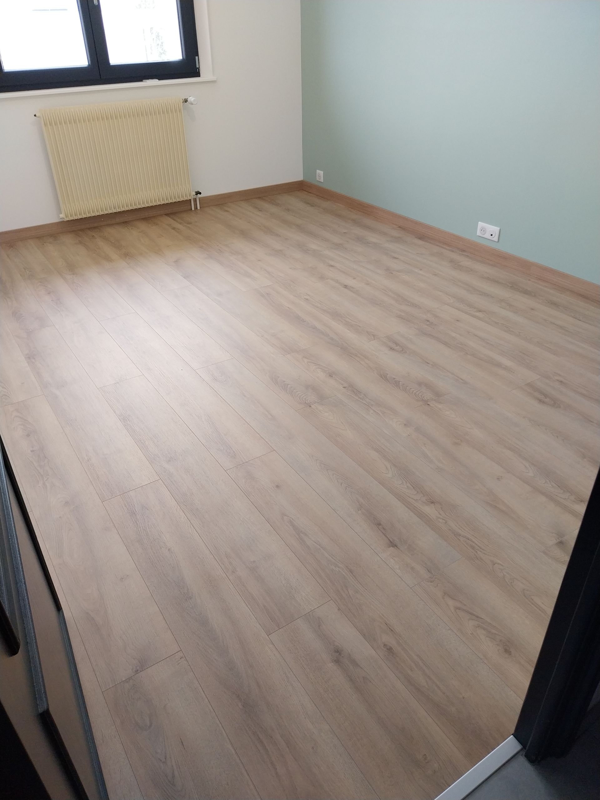 Pièce vide avec sol imitation bois et mur vert pâle. On aperçoit une fenêtre et un radiateur.