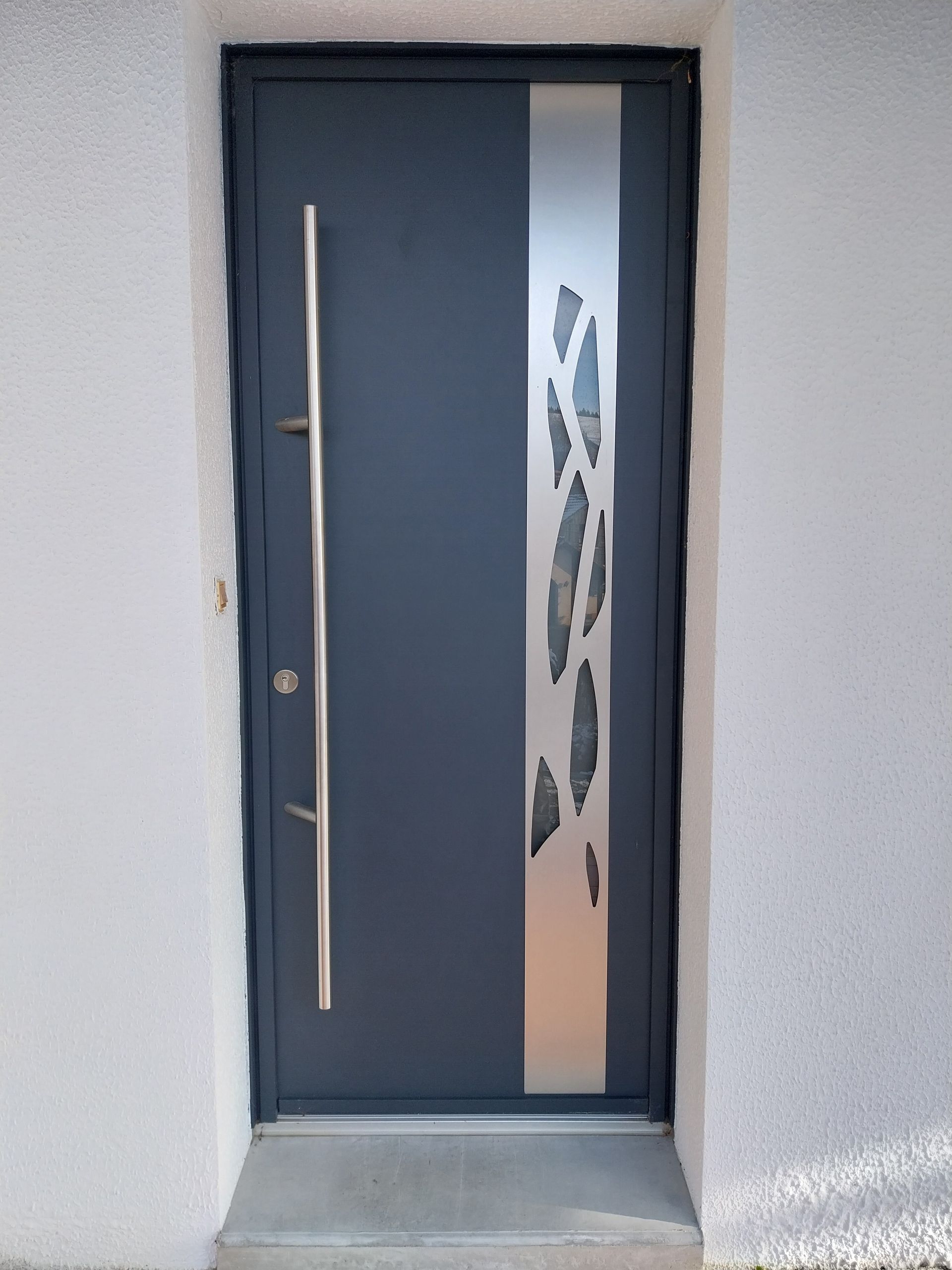 Porte d'entrée gris foncé avec un liseré argenté vertical et une longue poignée.