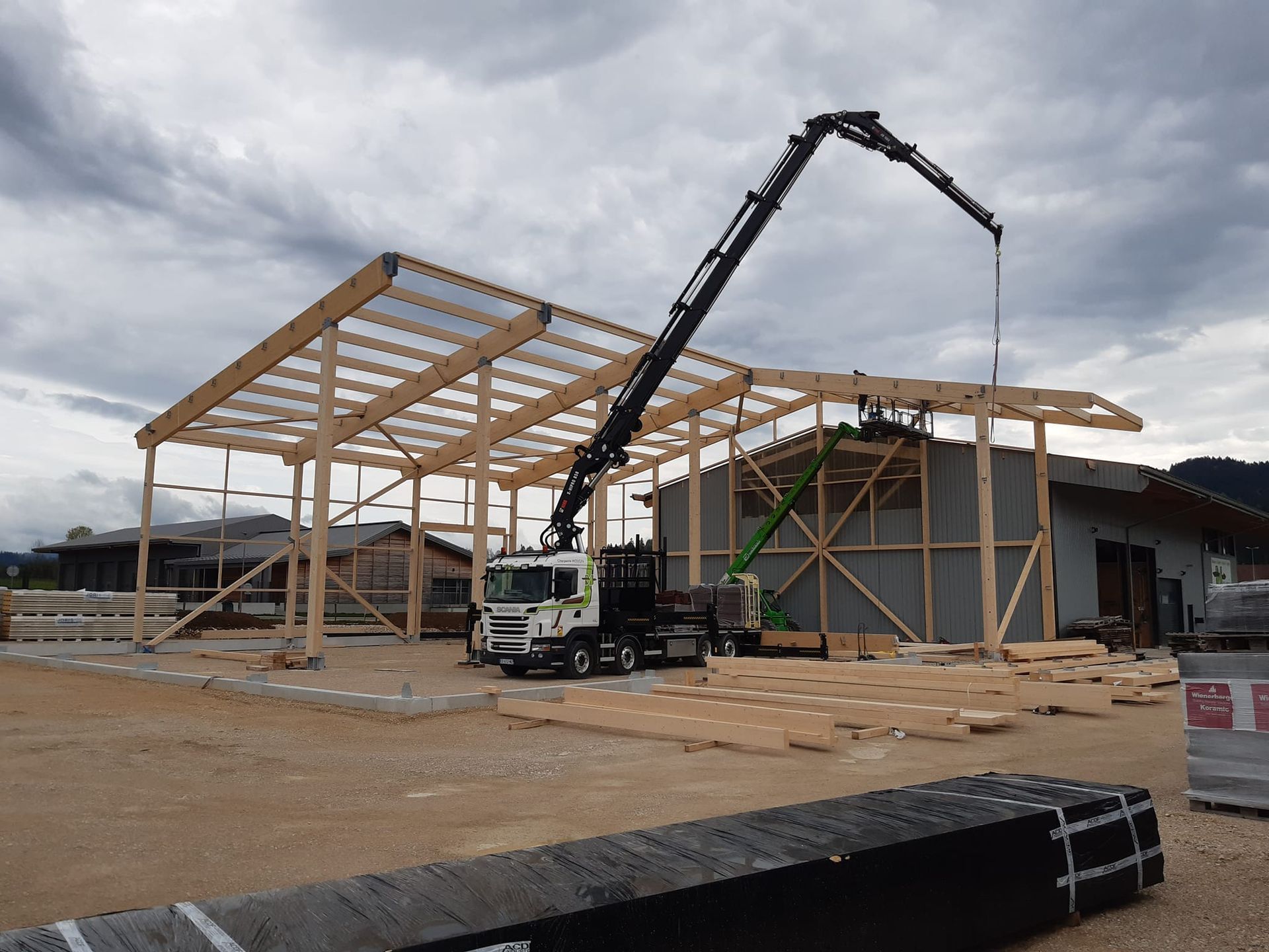 Chantier de construction avec grue soulevant des poutres de toiture en bois. Ciel nuageux.