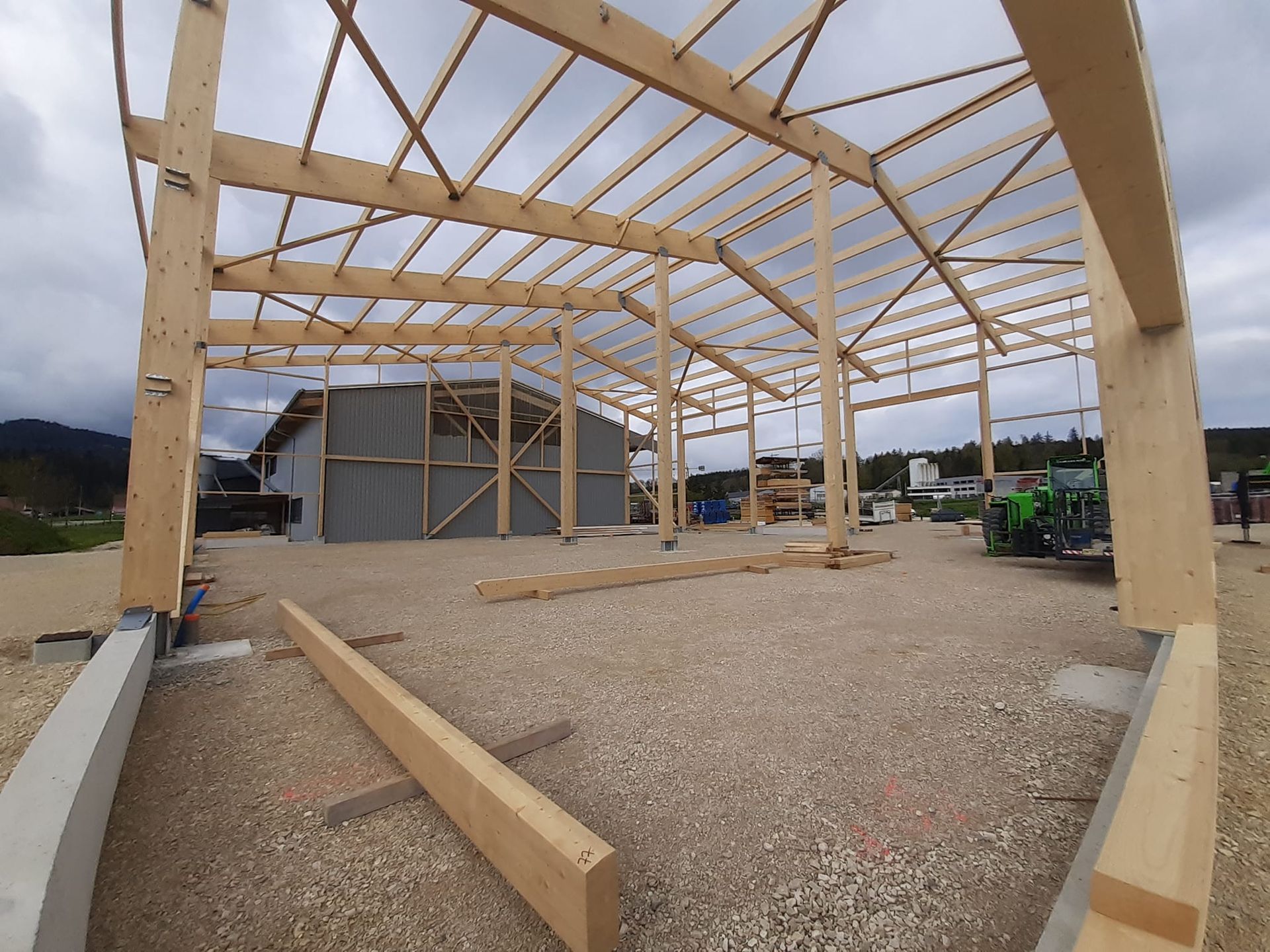 Bâtiment en bois en construction, vue intérieure avec poutres apparentes, sol en gravier, ciel couvert.