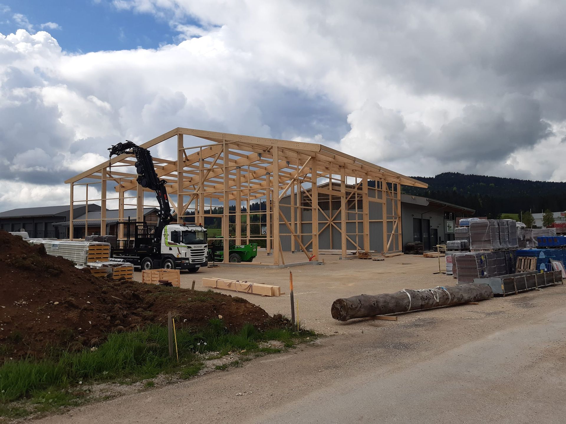 Construction d'une structure en bois à l'aide d'une grue sur un terrain gravillonné sous un ciel nuageux.