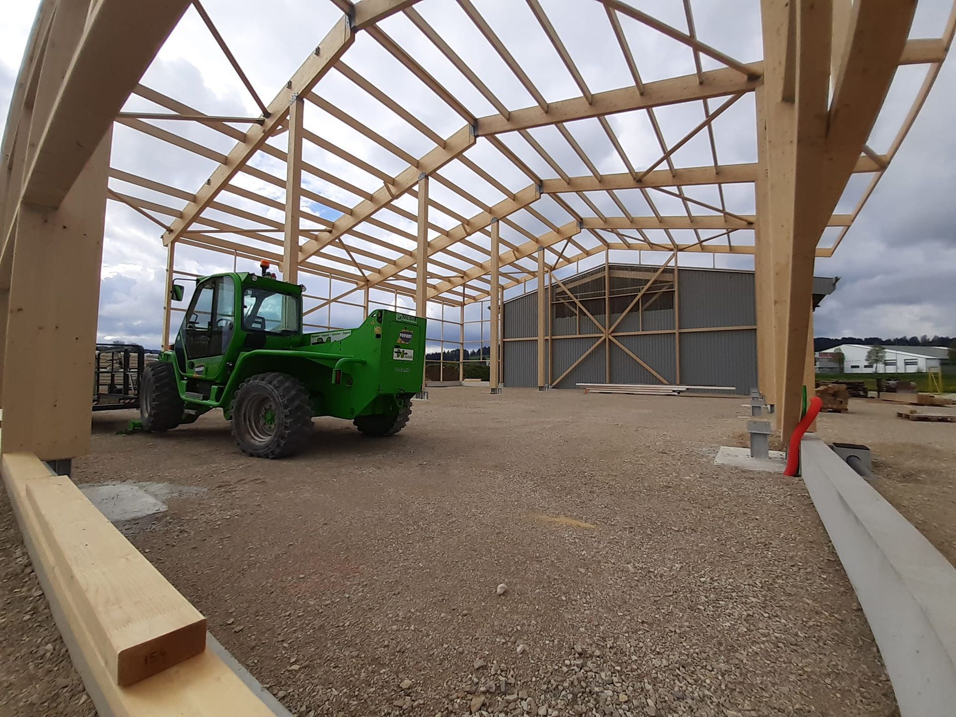 Charpente en bois d'un bâtiment en construction, sol en gravier, machines vertes, ciel nuageux.