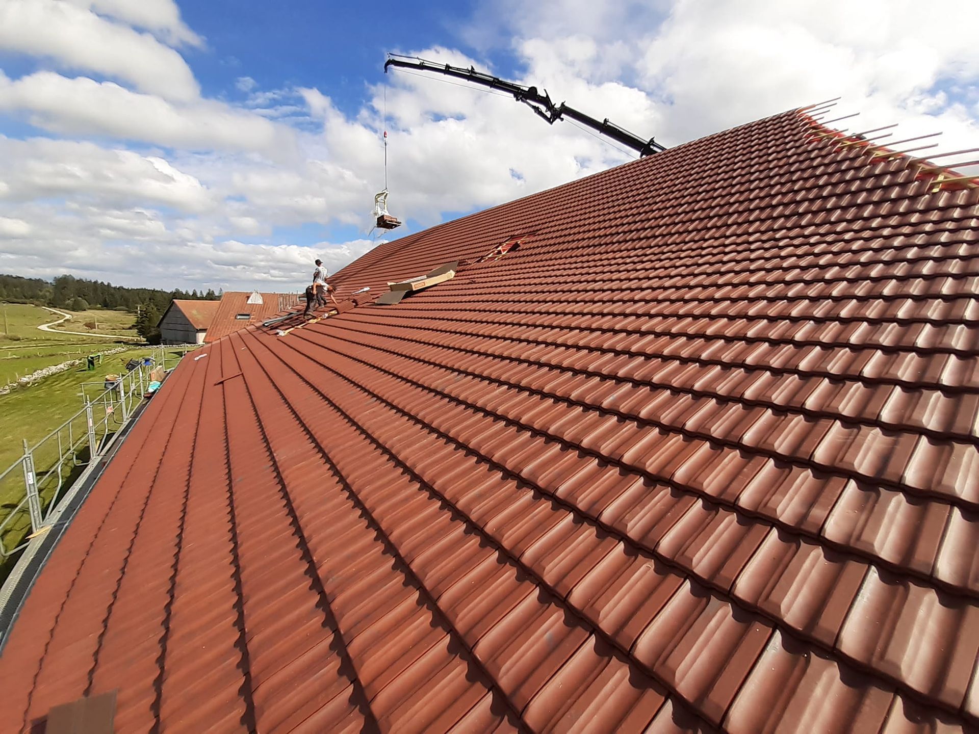 Toiture de tuiles rouges en cours de réfection, ciel bleu nuageux, grue soulevant des matériaux.