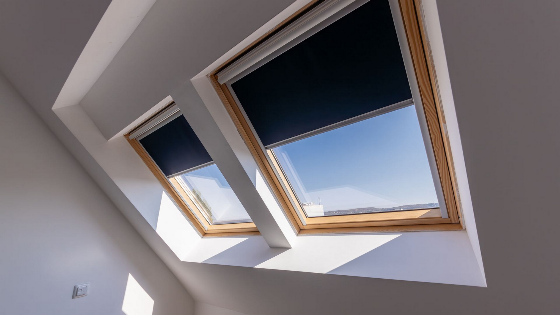 Deux fenêtres de toit aux stores bleu foncé dans une pièce blanche ; la lumière vive du soleil y pénètre.