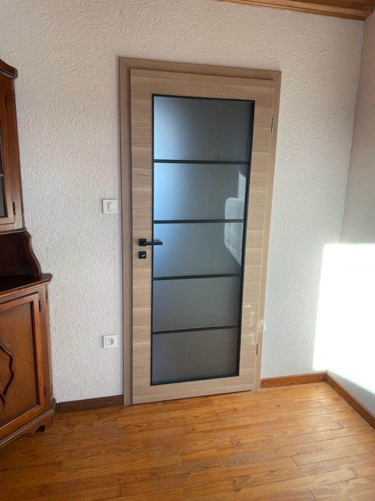 Porte en bois avec panneaux de verre dépoli, poignée noire et cadre beige. Installée dans une pièce avec parquet et murs blancs.