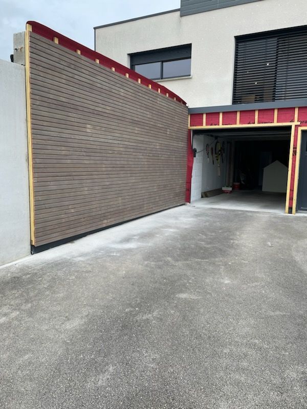 Un mur à panneaux bruns et à ossature rouge apparente borde l'entrée d'un garage. Cette structure contraste avec un bâtiment blanc moderne et l'asphalte.