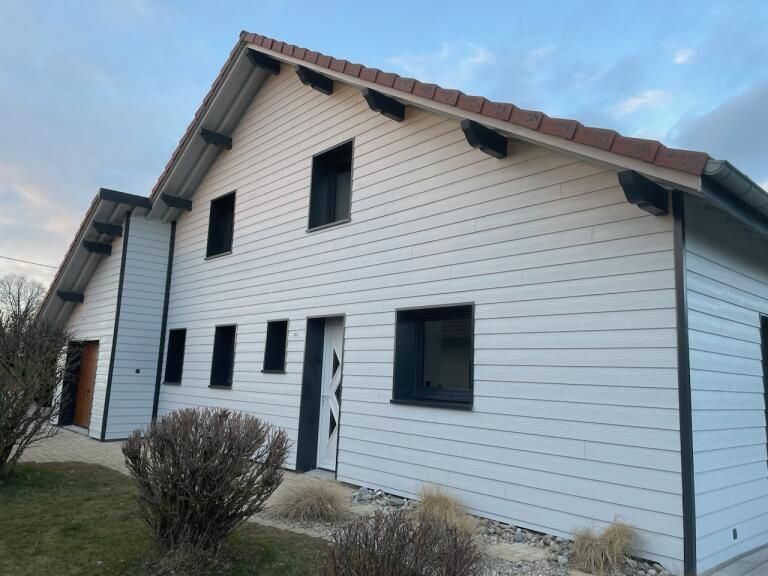 Maison blanche aux fenêtres et boiseries noires, toit rouge, ciel nuageux.