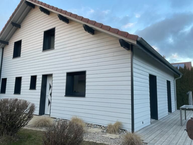 Maison blanche avec des boiseries et des fenêtres noires, un toit de tuiles rouges et une terrasse en bois.
