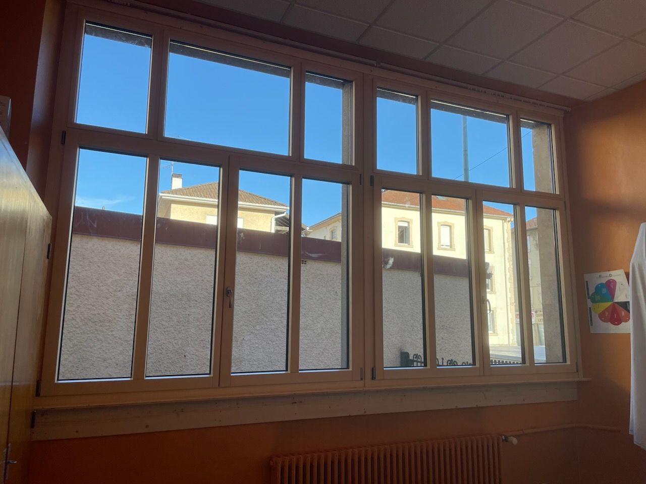 Fenêtres à ossature bois laissant apparaître un ciel bleu et le reflet des bâtiments environnants. Mur marron et radiateur en dessous.