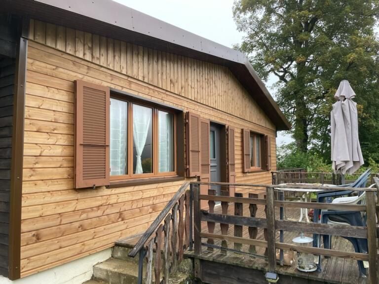 Cabane en bois avec finitions marron, volets et rambarde ; terrasse extérieure avec parasol.