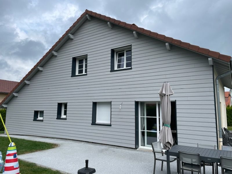 Maison grise avec bardage gris clair, encadrements de fenêtres foncés et toit rouge. Terrasse avec table et parasol.