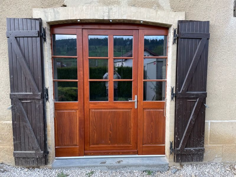 Porte en bois avec panneaux de verre et volets brun foncé, encadrée d'un cadre en pierre claire.