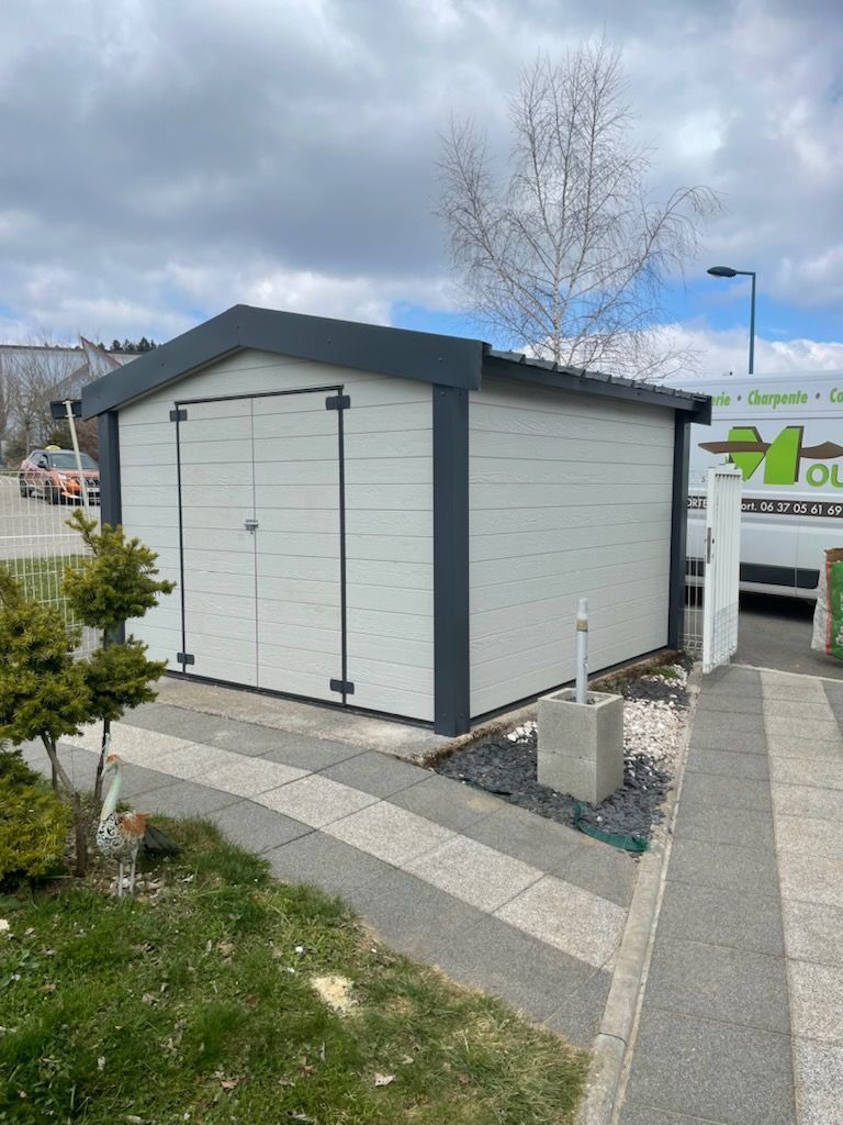 Abri de jardin gris et blanc avec des bordures gris foncé, devant un bâtiment entouré d'arbres et d'un trottoir.