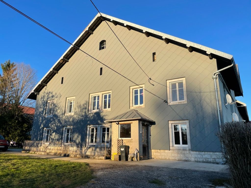 Maison grise avec de nombreuses fenêtres, un petit hall d'entrée et un toit sombre sous un ciel bleu.