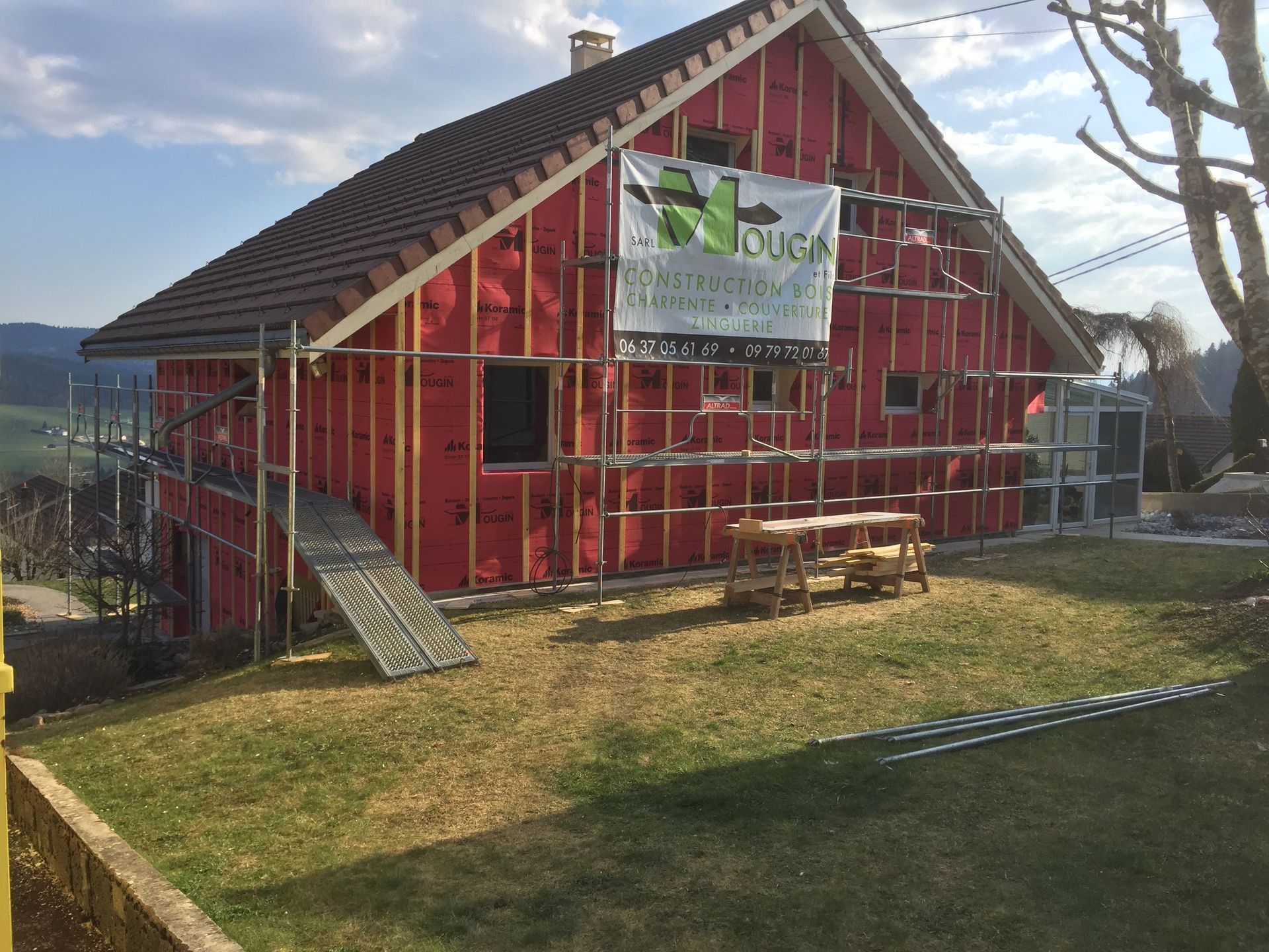 Maison en construction, bardage rouge, échafaudages, panneau, toit marron, pelouse, extérieur.