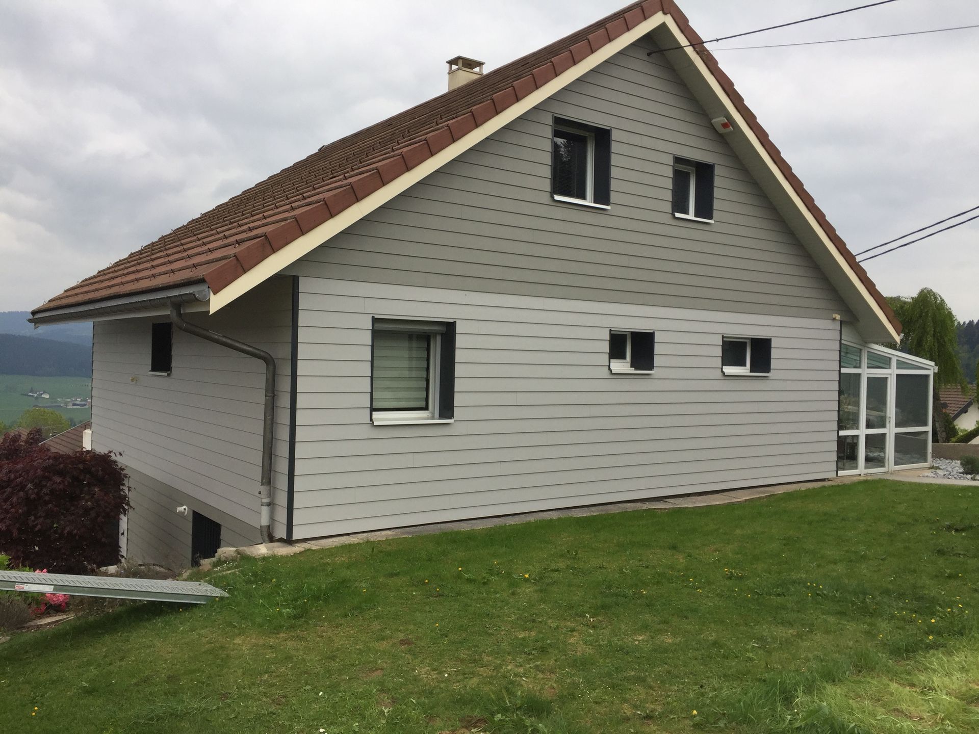 Maison grise au toit marron sur une colline herbeuse, avec fenêtres et petite véranda.