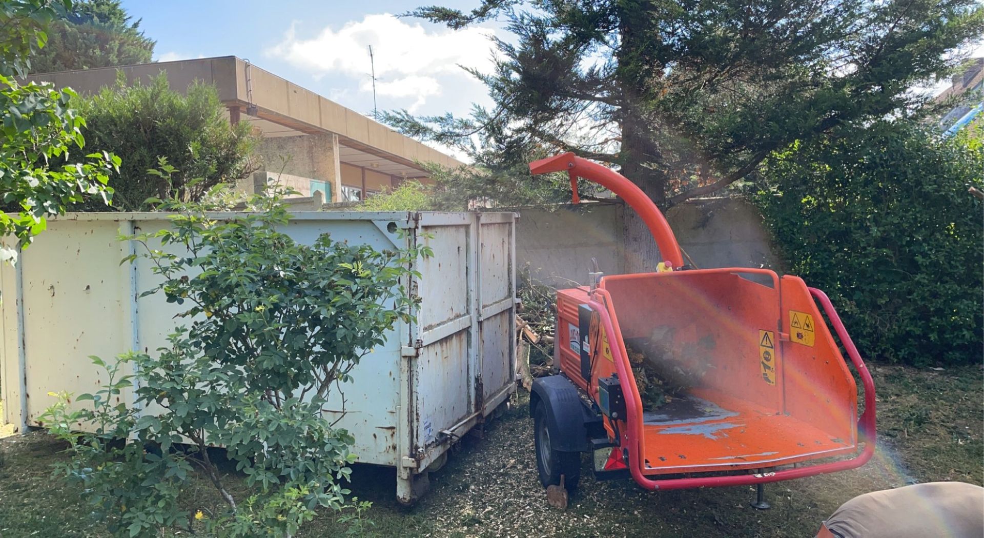 Broyeur de branches orange projetant des débris dans une grande benne à ordures, à l'extérieur.