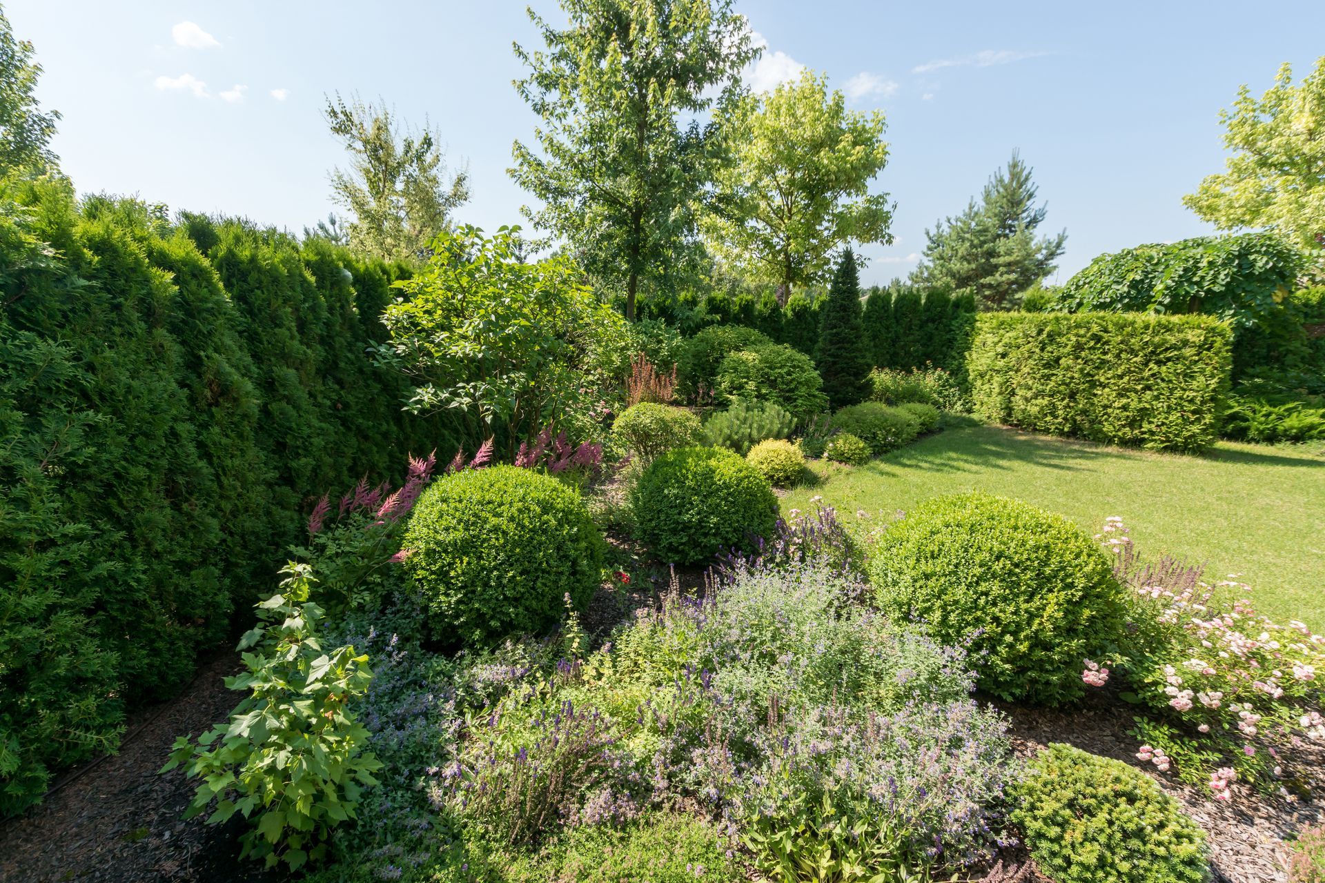 Un jardin luxuriant et ensoleillé, avec des haies taillées au cordeau, des pelouses verdoyantes et divers arbustes.