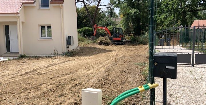 Une petite pelleteuse déblaye un terrain en terre battue jouxtant une maison beige.