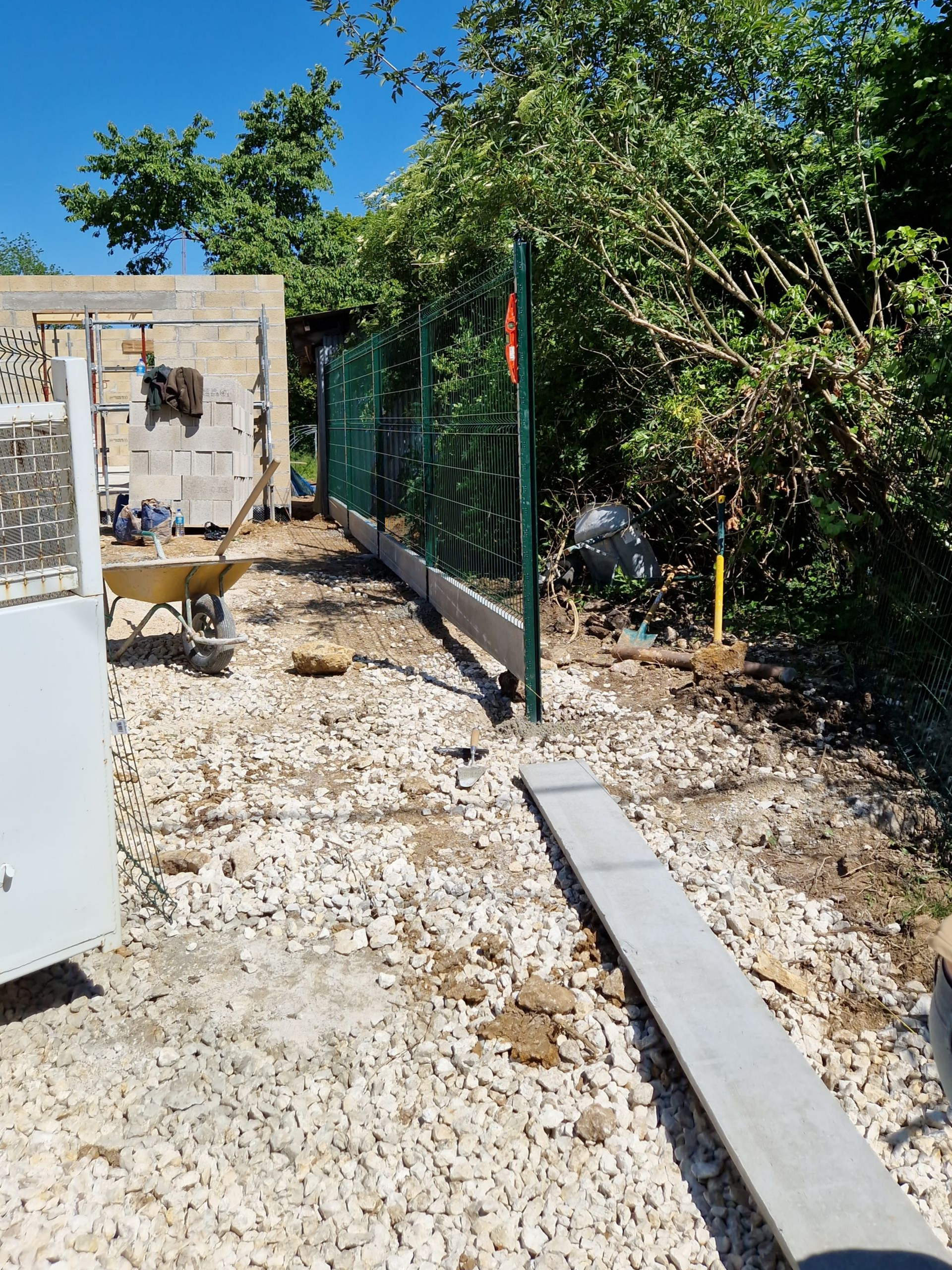 Chantier de construction avec une clôture en grillage partiellement construite et des dalles de béton sur gravier.