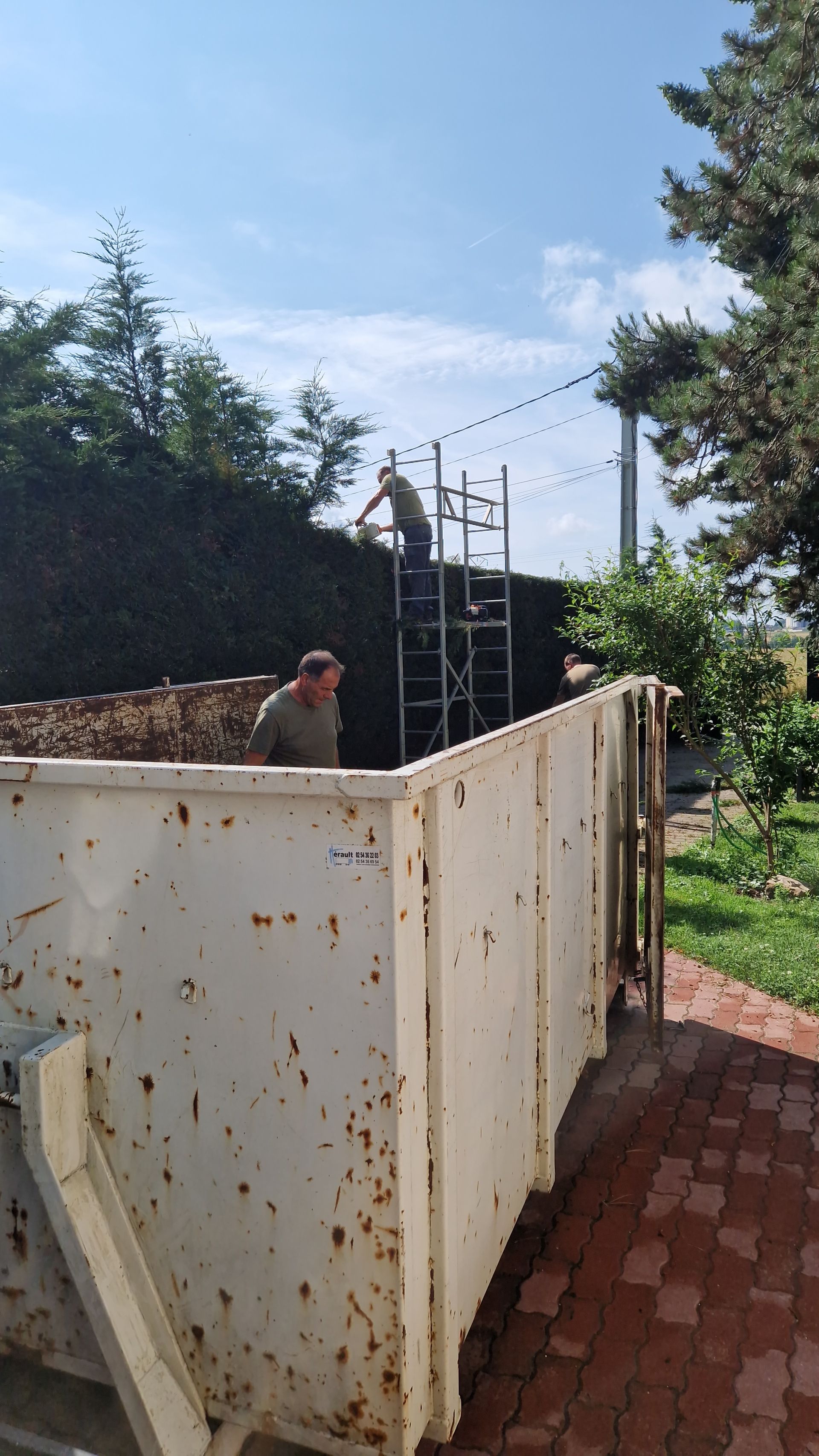 Deux personnes taillent une haie à côté d'une benne à ordures.