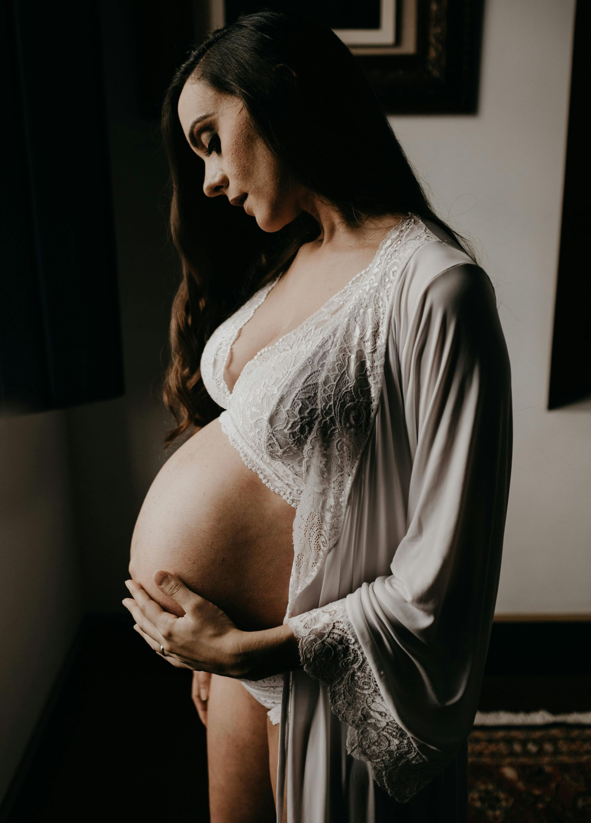 Schwangere Frau in weißer Lingerie streichelt ihren Bauch. Drinnen, in Fensternähe, sanftes Licht.