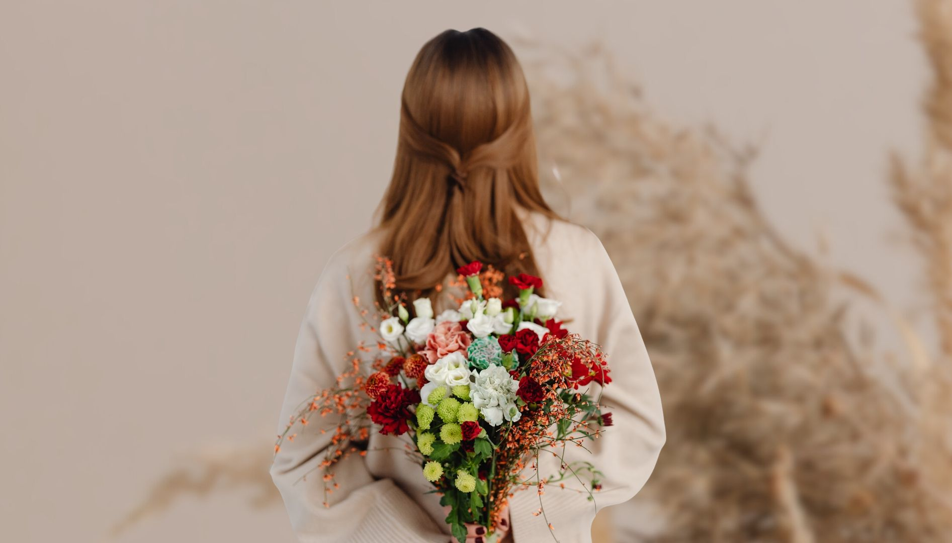 Frau hält einen bunten Blumenstrauß hinter dem Rücken. Beiger Pullover, heller Hintergrund.