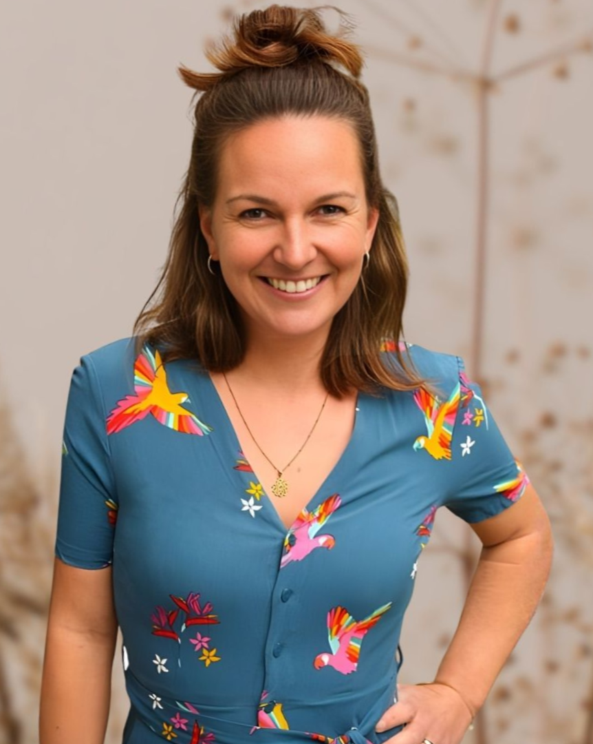 Frau lächelt, trägt ein blaues Kleid mit Vogelmuster, die Hand in die Hüfte gestemmt, das Haar zu einem Dutt gebunden.