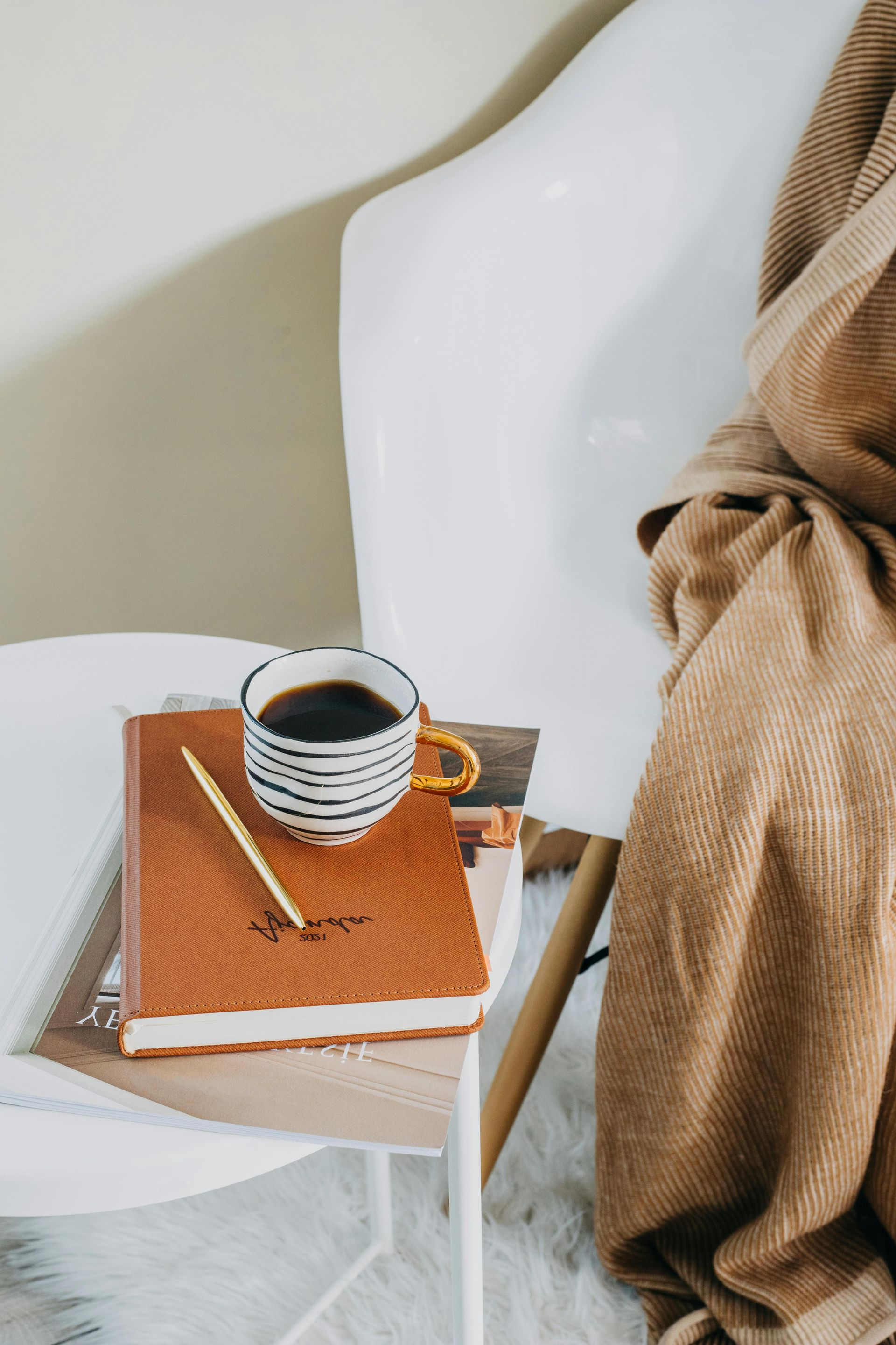 Kaffeetasse, Buch und Stift auf einem weißen Stuhl, seitlich darüber eine hellbraune Decke.