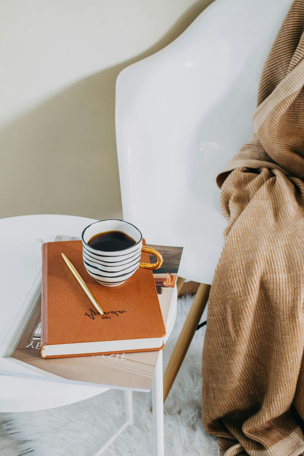 Kaffeetasse, Notizbuch und Stift auf einem weißen Stuhl, über den eine hellbraune Decke drapiert ist.