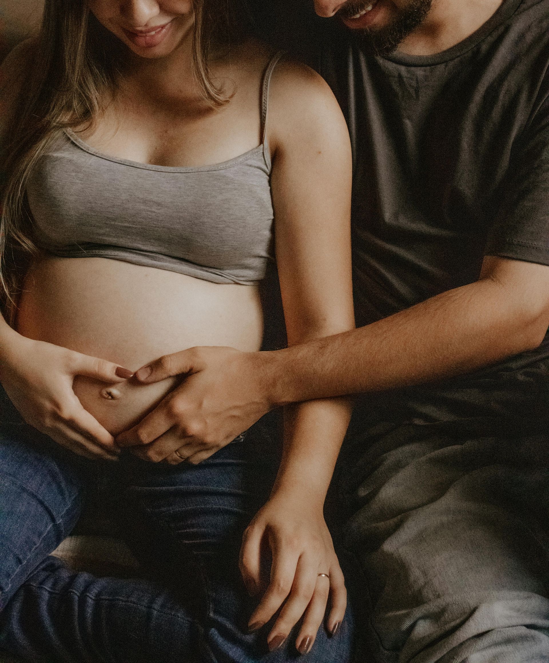 Schwangere Frau in grauem Top und Jeans, die Hände eines Mannes formen ein Herz auf ihrem Bauch.