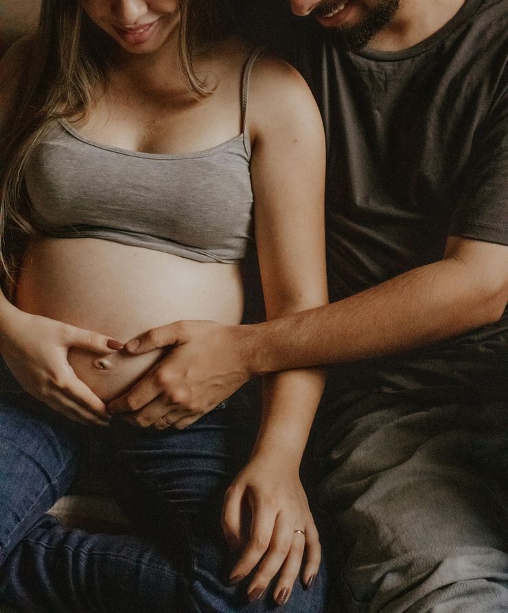 Schwangere Frau in grauem Top und Jeans, die Hände eines Mannes formen ein Herz auf ihrem Bauch.