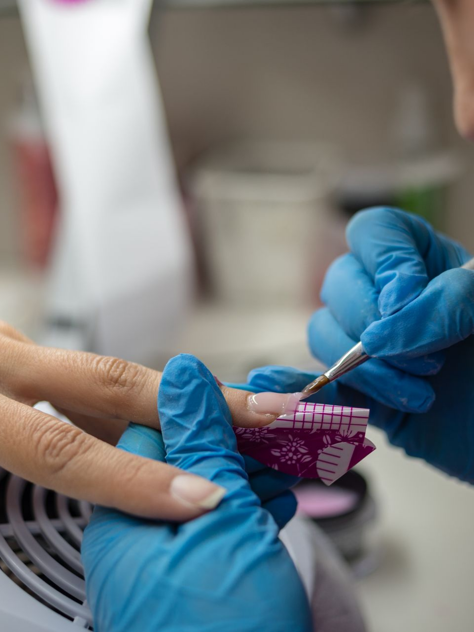 Une personne portant des gants bleus applique du vernis à ongles sur l'ongle d'un client à l'aide d'un pinceau et d'un moule à ongles dans un salon de coiffure.